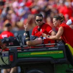 FootballR - NFL - Ein verletzter Sportler, Grayson McCall, liegt auf einer Trage in einem grünen John Deere-Fahrzeug, begleitet von medizinischem Personal in roten Hemden. McCall trägt eine schwarze Uniform und streckt zwei Finger in die Höhe, während er von einer Menschenmenge in roten Sitzen umgeben ist, was auf eine Sportveranstaltung hindeutet. Diese Beschreibung wurde automatisch generiert.