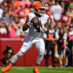 FootballR - NFL - Deshaun Watson in weißer Uniform mit orangefarbenen und braunen Akzenten hält einen Football und bereitet sich zum Wurf vor. Er steht auf einem grünen Feld mit einer verschwommenen Menschenmenge im Hintergrund. Er trägt einen orangefarbenen Helm und weiße Stollenschuhe, was darauf hindeutet, dass gerade ein Spiel läuft. Diese Beschreibung wurde automatisch generiert.