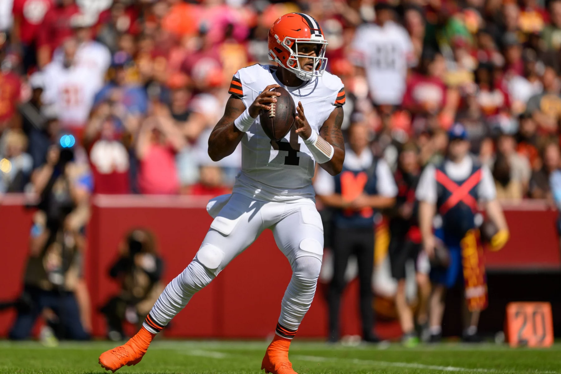 FootballR - NFL - Deshaun Watson in weißer Uniform mit orangefarbenen und braunen Akzenten hält einen Football und bereitet sich zum Wurf vor. Er steht auf einem grünen Feld mit einer verschwommenen Menschenmenge im Hintergrund. Er trägt einen orangefarbenen Helm und weiße Stollenschuhe, was darauf hindeutet, dass gerade ein Spiel läuft. Diese Beschreibung wurde automatisch generiert.