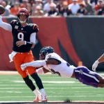 FootballR - NFL - Ein Footballspieler, Joe Burrow, in einem orange-schwarzen Trikot der Cincinnati Bengals, Nummer 9, wirft einen Pass, während ein anderer Spieler in einem weiß-lila Trikot der Baltimore Ravens auf ihn zuspringt. Das Spiel findet auf dem grünen Feld statt, im Hintergrund halten verschwommene Zuschauer den spannenden Moment fest. Diese Beschreibung wurde automatisch generiert.