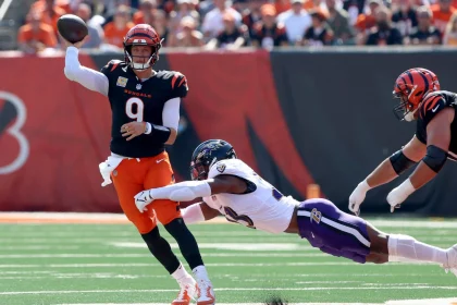 FootballR - NFL - Ein Footballspieler, Joe Burrow, in einem orange-schwarzen Trikot der Cincinnati Bengals, Nummer 9, wirft einen Pass, während ein anderer Spieler in einem weiß-lila Trikot der Baltimore Ravens auf ihn zuspringt. Das Spiel findet auf dem grünen Feld statt, im Hintergrund halten verschwommene Zuschauer den spannenden Moment fest. Diese Beschreibung wurde automatisch generiert.