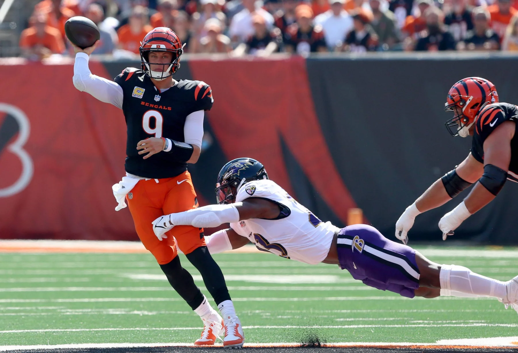 FootballR - NFL - Ein Footballspieler, Joe Burrow, in einem orange-schwarzen Trikot der Cincinnati Bengals, Nummer 9, wirft einen Pass, während ein anderer Spieler in einem weiß-lila Trikot der Baltimore Ravens auf ihn zuspringt. Das Spiel findet auf dem grünen Feld statt, im Hintergrund halten verschwommene Zuschauer den spannenden Moment fest. Diese Beschreibung wurde automatisch generiert.