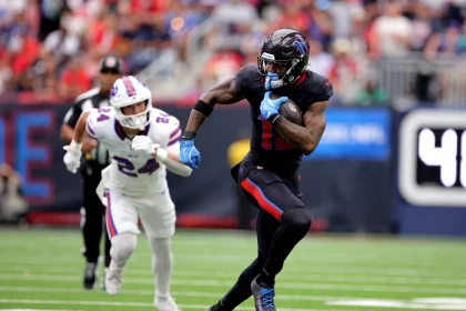 FootballR - NFL - Nico Collins in schwarzer Uniform rennt mit dem Ball über das Feld, verfolgt von einem anderen Spieler in weißer Uniform mit roten und blauen Akzenten. Diese Beschreibung wurde automatisch generiert.