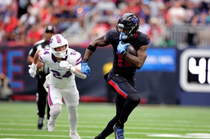 FootballR - NFL - Nico Collins in schwarzer Uniform rennt mit dem Ball über das Feld, verfolgt von einem anderen Spieler in weißer Uniform mit roten und blauen Akzenten. Diese Beschreibung wurde automatisch generiert.