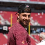 FootballR - NFL - Ein Mann mit Bart, Ricky Pearsall, trägt ein rotes Hemd und eine schwarze Kappe mit einem Sportlogo. Er lächelt, während er in einem Stadion steht und nach hinten blickt. Die Tribünen hinter ihm sind größtenteils leer, nur wenige Zuschauer sind da. Die Sonne scheint hell über das im Hintergrund sichtbare Fußballfeld. Diese Beschreibung wurde automatisch generiert.