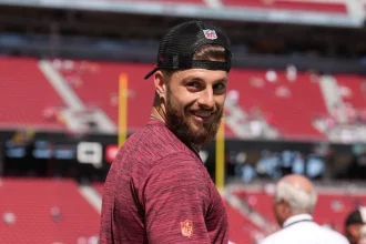 FootballR - NFL - Ein Mann mit Bart, Ricky Pearsall, trägt ein rotes Hemd und eine schwarze Kappe mit einem Sportlogo. Er lächelt, während er in einem Stadion steht und nach hinten blickt. Die Tribünen hinter ihm sind größtenteils leer, nur wenige Zuschauer sind da. Die Sonne scheint hell über das im Hintergrund sichtbare Fußballfeld. Diese Beschreibung wurde automatisch generiert.