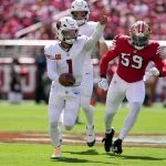 FootballR - NFL - In einem spannenden Showdown, der als „Sensation in Santa Clara“ bezeichnet wird, stürmt ein Footballspieler in weißer Uniform mit dem Ball in der Hand - Kyler Murray - nach vorne, verfolgt von einem Gegner in Rot. Während Jubel aus dem voll besetzten Stadion ausbricht, glitzern die Teamlogos während dieses intensiven Aufeinandertreffens der Cardinals gegen die 49ers in der Sonne. Diese Beschreibung wurde automatisch generiert.