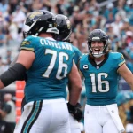 FootballR - NFL - Footballspieler in blaugrünen Trikots, darunter Trevor Lawrence, feiern während eines Spiels auf dem Feld. Nummer 16, mit einem blaugrünen Visier auf seinem Helm, lächelt aufgeregt. Nummer 76 steht in der Nähe und blickt ihm entgegen. Im Hintergrund sind verschwommene Zuschauer in einem lebhaften Stadion zu sehen. Diese Beschreibung wurde automatisch generiert.