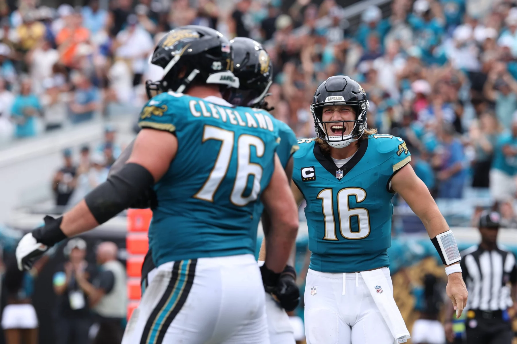 FootballR - NFL - Footballspieler in blaugrünen Trikots, darunter Trevor Lawrence, feiern während eines Spiels auf dem Feld. Nummer 16, mit einem blaugrünen Visier auf seinem Helm, lächelt aufgeregt. Nummer 76 steht in der Nähe und blickt ihm entgegen. Im Hintergrund sind verschwommene Zuschauer in einem lebhaften Stadion zu sehen. Diese Beschreibung wurde automatisch generiert.