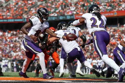 FootballR - AFC North - NFL Offensiv-Spektakel - Der Quarterback der Baltimore Ravens - Lamar Jackson - im weißen Trikot übergibt den Football während eines AFC North Spiels gegen die Bengals an einen Running Back. Der Running Back, ebenfalls im weißen Trikot, bereitet sich auf seinen Vorstoß vor, während Abwehr- und Angriffsspieler beider Teams, darunter auch solche in orangefarbenen Trikots, in Aktion treten. Diese Beschreibung wurde automatisch generiert.