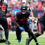 FootballR - NFL - Cam Akers, in schwarzer Uniform mit blauen Handschuhen, sprintet während eines spannenden Spiels mit dem Ball über das Feld. Teamkollegen und Gegner umringen ihn auf dem leuchtend grünen Feld. Im unscharfen Hintergrund füllen Zuschauer die Tribünen und verfolgen gespannt jede Bewegung. Diese Beschreibung wurde automatisch generiert.