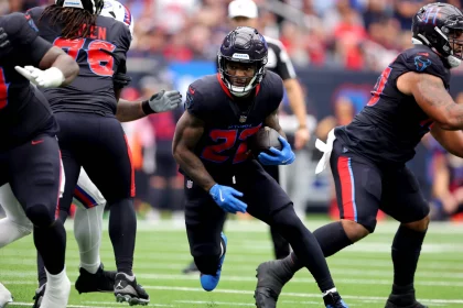 FootballR - NFL - Cam Akers, in schwarzer Uniform mit blauen Handschuhen, sprintet während eines spannenden Spiels mit dem Ball über das Feld. Teamkollegen und Gegner umringen ihn auf dem leuchtend grünen Feld. Im unscharfen Hintergrund füllen Zuschauer die Tribünen und verfolgen gespannt jede Bewegung. Diese Beschreibung wurde automatisch generiert.