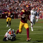 FootballR - NFL Week 5 Analyse - Während der 5. NFL-Woche weicht ein Footballspieler in kastanienbrauner und gelber Uniform - Jayden Daniels - gekonnt einem Tackle eines Gegners in Weiß und Orange aus. Inmitten des sonnenbeschienenen Stadions bilden andere Spieler und eine lebhafte Menge die Kulisse dieses intensiven Spielmoments. Diese Beschreibung wurde automatisch generiert.