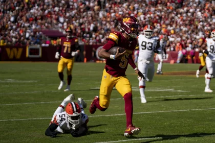 FootballR - NFL Week 5 Analyse - Während der 5. NFL-Woche weicht ein Footballspieler in kastanienbrauner und gelber Uniform - Jayden Daniels - gekonnt einem Tackle eines Gegners in Weiß und Orange aus. Inmitten des sonnenbeschienenen Stadions bilden andere Spieler und eine lebhafte Menge die Kulisse dieses intensiven Spielmoments. Diese Beschreibung wurde automatisch generiert.