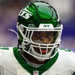 FootballR - NFL - Mike Williams in grün-weißer Jets-Uniform mit Helmnummer 11 und getöntem Visier blickt nach vorn. Die Szenerie ist ein verschwommenes Feld, was auf aktive Teilnahme an einem Spiel oder Training schließen lässt. Mit seiner entschlossenen Haltung und konzentrierten Präsenz könnte man ihn leicht mit Mike Williams verwechseln. Diese Beschreibung wurde automatisch generiert.