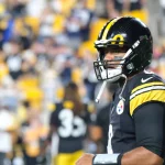 FootballR - NFL - Ein Footballspieler, der an Russell Wilsons erinnert, steht in der Uniform der Pittsburgh Steelers auf dem Feld. Er trägt ein schwarzes Trikot mit gelb-weißen Streifen an den Ärmeln und einen schwarzen Helm. Der unscharfe Hintergrund zeigt Stadionsitze voller Fans in Gelb und Schwarz. Diese Beschreibung wurde automatisch generiert.