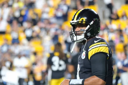 FootballR - NFL - Ein Footballspieler, der an Russell Wilsons erinnert, steht in der Uniform der Pittsburgh Steelers auf dem Feld. Er trägt ein schwarzes Trikot mit gelb-weißen Streifen an den Ärmeln und einen schwarzen Helm. Der unscharfe Hintergrund zeigt Stadionsitze voller Fans in Gelb und Schwarz. Diese Beschreibung wurde automatisch generiert.