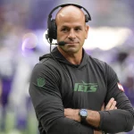 FootballR - NFL - Ein kahlköpfiger Mann mit Headset steht auf einem Footballfeld. Er trägt einen schwarzen Kapuzenpullover mit dem „Jets“-Logo und verschränkt die Arme. Es ist Robert Saleh, Cheftrainer der New York Jets. Im unscharfen Hintergrund sind Spieler in weißen und violetten Uniformen zu sehen. An seinem Handgelenk ist eine Uhr zu sehen. Diese Beschreibung wurde automatisch generiert.