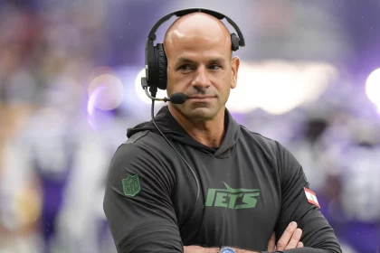 FootballR - NFL - Ein kahlköpfiger Mann mit Headset steht auf einem Footballfeld. Er trägt einen schwarzen Kapuzenpullover mit dem „Jets“-Logo und verschränkt die Arme. Es ist Robert Saleh, Cheftrainer der New York Jets. Im unscharfen Hintergrund sind Spieler in weißen und violetten Uniformen zu sehen. An seinem Handgelenk ist eine Uhr zu sehen. Diese Beschreibung wurde automatisch generiert.