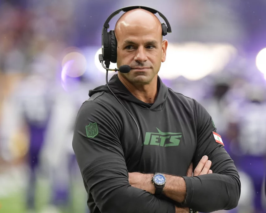 FootballR - NFL - Ein kahlköpfiger Mann mit Headset steht auf einem Footballfeld. Er trägt einen schwarzen Kapuzenpullover mit dem „Jets“-Logo und verschränkt die Arme. Es ist Robert Saleh, Cheftrainer der New York Jets. Im unscharfen Hintergrund sind Spieler in weißen und violetten Uniformen zu sehen. An seinem Handgelenk ist eine Uhr zu sehen. Diese Beschreibung wurde automatisch generiert.