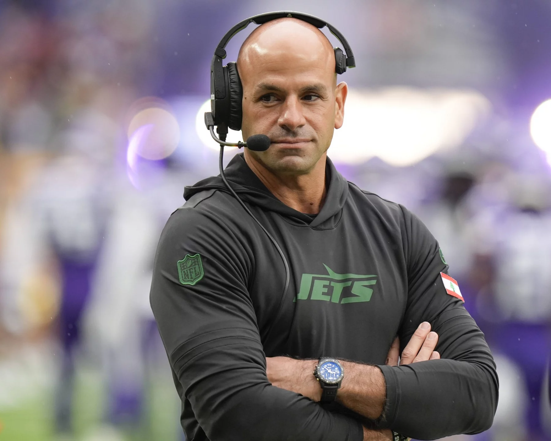 FootballR - NFL - Ein kahlköpfiger Mann mit Headset steht auf einem Footballfeld. Er trägt einen schwarzen Kapuzenpullover mit dem „Jets“-Logo und verschränkt die Arme. Es ist Robert Saleh, Cheftrainer der New York Jets. Im unscharfen Hintergrund sind Spieler in weißen und violetten Uniformen zu sehen. An seinem Handgelenk ist eine Uhr zu sehen. Diese Beschreibung wurde automatisch generiert.