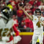 FootballR - NFL - Derek Carr, in weißer Uniform, bereitet sich darauf vor, den Ball zu werfen, wobei er seinen rechten Arm nach vorne streckt. Ein weiterer Spieler in einem weißen Trikot läuft im Vordergrund vor einer verschwommenen, rot gekleideten Menge. Die Szene fängt die Intensität eines laufenden Spiels ein. Diese Beschreibung wurde automatisch generiert.