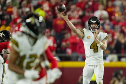 FootballR - NFL - Derek Carr, in weißer Uniform, bereitet sich darauf vor, den Ball zu werfen, wobei er seinen rechten Arm nach vorne streckt. Ein weiterer Spieler in einem weißen Trikot läuft im Vordergrund vor einer verschwommenen, rot gekleideten Menge. Die Szene fängt die Intensität eines laufenden Spiels ein. Diese Beschreibung wurde automatisch generiert.