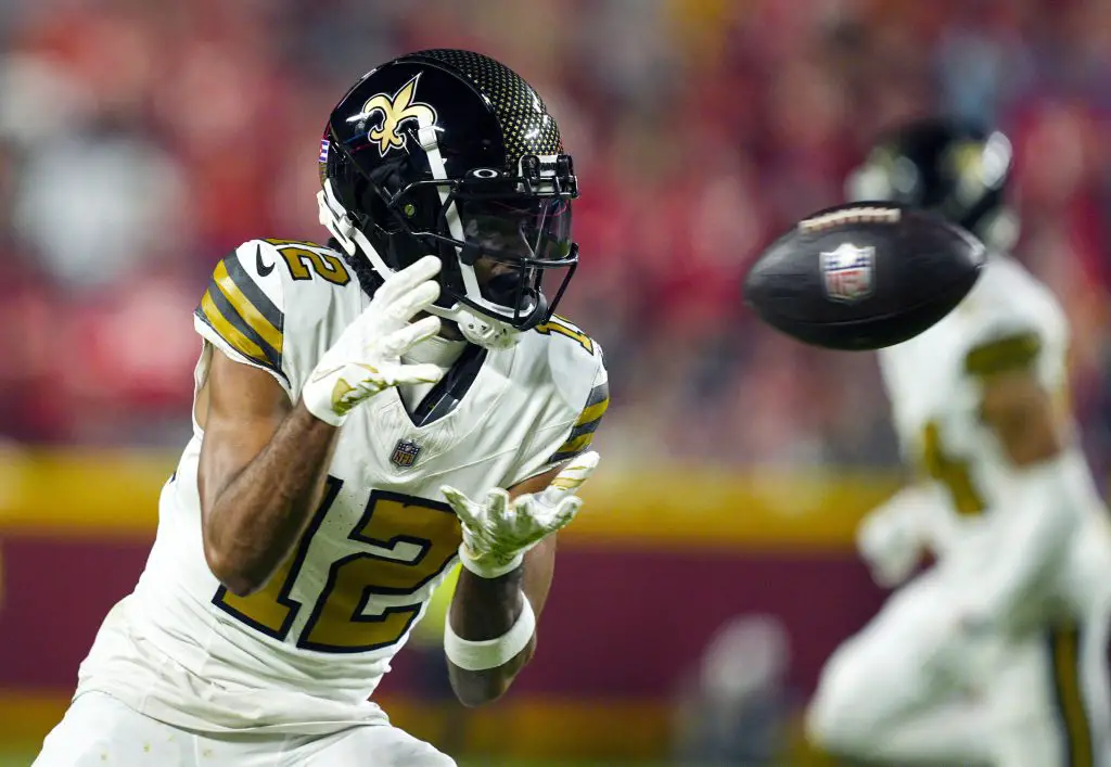 FootballR - NFL - Ein Footballspieler, der an Chris Olave erinnert, trägt eine weiß-schwarze Uniform der New Orleans Saints mit der Nummer 12. Er konzentriert sich voll und ganz auf das anstehende Spiel und trägt einen schwarzen Helm mit dem Logo des Teams. Im Hintergrund feuert die verschwommene Menge im Stadion ihn an. Diese Beschreibung wurde automatisch generiert.