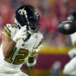 FootballR - NFL - Ein Footballspieler, der an Chris Olave erinnert, trägt eine weiß-schwarze Uniform der New Orleans Saints mit der Nummer 12. Er konzentriert sich voll und ganz auf das anstehende Spiel und trägt einen schwarzen Helm mit dem Logo des Teams. Im Hintergrund feuert die verschwommene Menge im Stadion ihn an. Diese Beschreibung wurde automatisch generiert.