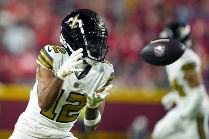 FootballR - NFL - Ein Footballspieler, der an Chris Olave erinnert, trägt eine weiß-schwarze Uniform der New Orleans Saints mit der Nummer 12. Er konzentriert sich voll und ganz auf das anstehende Spiel und trägt einen schwarzen Helm mit dem Logo des Teams. Im Hintergrund feuert die verschwommene Menge im Stadion ihn an. Diese Beschreibung wurde automatisch generiert.