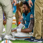 FootballR - NFL - Ein Footballspieler in einem blaugrünen Trikot mit einer weißen Nummer, der Tua Tagovailoa ähnelt, sitzt auf dem Feld, umgeben von medizinischem Personal in Khakihosen. Der Spieler scheint verletzt zu sein, er trägt eine Schiene an seinem rechten Handgelenk und einen Eisbeutel auf seinem linken Knie. Das Personal kümmert sich um ihn, während er sichtlich verstört aussieht. Diese Beschreibung wurde automatisch generiert.