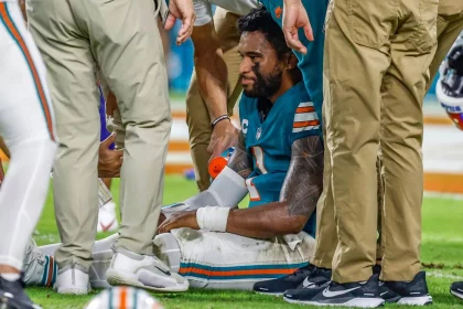 FootballR - NFL - Ein Footballspieler in einem blaugrünen Trikot mit einer weißen Nummer, der Tua Tagovailoa ähnelt, sitzt auf dem Feld, umgeben von medizinischem Personal in Khakihosen. Der Spieler scheint verletzt zu sein, er trägt eine Schiene an seinem rechten Handgelenk und einen Eisbeutel auf seinem linken Knie. Das Personal kümmert sich um ihn, während er sichtlich verstört aussieht. Diese Beschreibung wurde automatisch generiert.