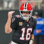 FootballR - NFL Fantasy Football - Kirk Cousins in schwarzer Uniform mit der Nummer 18 gestikuliert während eines Spiels. Er trägt einen roten Helm mit einem schwarzen Streifen. Im Hintergrund ist das Spielfeld verschwommen, ein weiterer Spieler in Blau ist zu sehen. Die Szene fängt Action und Konzentration ein. Diese Beschreibung wurde automatisch generiert.