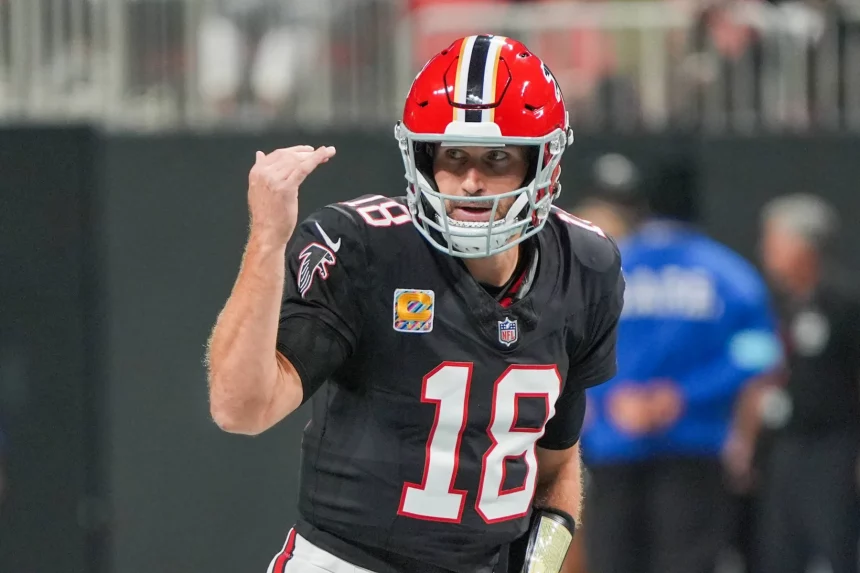 FootballR - NFL Fantasy Football - Kirk Cousins in schwarzer Uniform mit der Nummer 18 gestikuliert während eines Spiels. Er trägt einen roten Helm mit einem schwarzen Streifen. Im Hintergrund ist das Spielfeld verschwommen, ein weiterer Spieler in Blau ist zu sehen. Die Szene fängt Action und Konzentration ein. Diese Beschreibung wurde automatisch generiert.