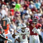 FootballR - College Football - Inmitten der lebhaften Atmosphäre eines College-Footballspiels wirft ein Spieler im roten Trikot mit der Nummer 4 gekonnt einen Pass. Gegner in weißen Trikots, einer davon mit der Nummer 6, nähern sich rasch. Im Hintergrund summt es von verschwommenen Zuschauern, während das Sonnenlicht die aufregende Action auf dem Spielfeld hervorhebt. Diese Beschreibung wurde automatisch generiert.