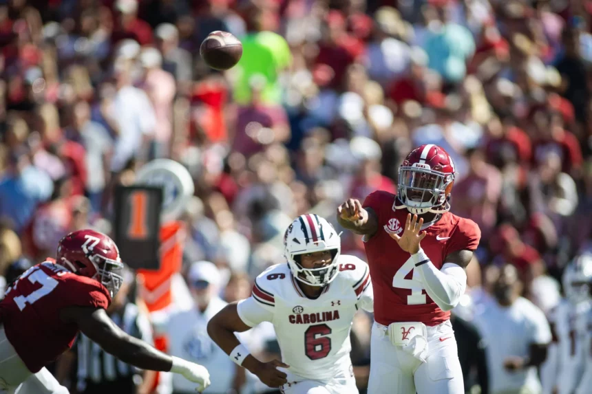 FootballR - College Football - Inmitten der lebhaften Atmosphäre eines College-Footballspiels wirft ein Spieler im roten Trikot mit der Nummer 4 gekonnt einen Pass. Gegner in weißen Trikots, einer davon mit der Nummer 6, nähern sich rasch. Im Hintergrund summt es von verschwommenen Zuschauern, während das Sonnenlicht die aufregende Action auf dem Spielfeld hervorhebt. Diese Beschreibung wurde automatisch generiert.