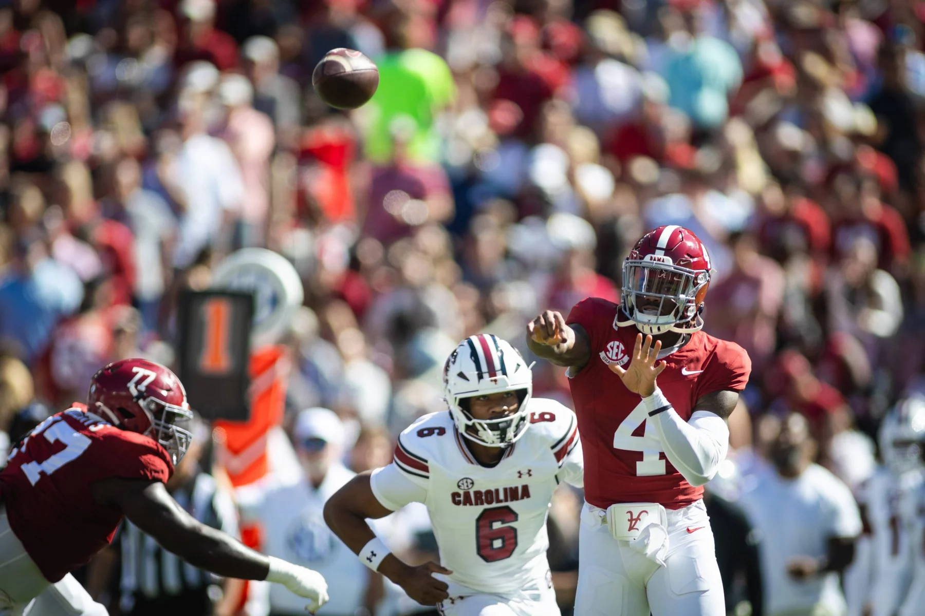 FootballR - College Football - Inmitten der lebhaften Atmosphäre eines College-Footballspiels wirft ein Spieler im roten Trikot mit der Nummer 4 gekonnt einen Pass. Gegner in weißen Trikots, einer davon mit der Nummer 6, nähern sich rasch. Im Hintergrund summt es von verschwommenen Zuschauern, während das Sonnenlicht die aufregende Action auf dem Spielfeld hervorhebt. Diese Beschreibung wurde automatisch generiert.