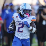 FootballR - NFL - Ein College Football Spieler in einem weißen Trikot mit der Nummer 2 und „BOISE STATE“, Ashton Jeanty, rennt mit einem Football in der Hand über das Feld. Auf seinem weißen Helm ist ein blaues Pferdelogo zu sehen, ergänzt durch blaue Hosen und rosa Ausrüstung. Begeisterte Fans und Offizielle sorgen für eine lebendige Kulisse. Diese Beschreibung wurde automatisch generiert.