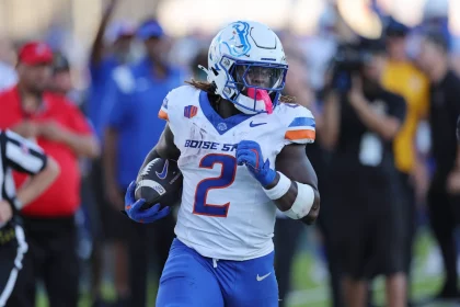 FootballR - NFL - Ein College Football Spieler in einem weißen Trikot mit der Nummer 2 und „BOISE STATE“, Ashton Jeanty, rennt mit einem Football in der Hand über das Feld. Auf seinem weißen Helm ist ein blaues Pferdelogo zu sehen, ergänzt durch blaue Hosen und rosa Ausrüstung. Begeisterte Fans und Offizielle sorgen für eine lebendige Kulisse. Diese Beschreibung wurde automatisch generiert.