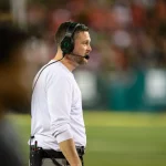 FootballR - NCAA College Football - Ein Trainer, Dan Lanning, in einem weißen Langarmshirt mit Kopfhörern steht am Spielfeldrand eines NCAA-Showdowns. Die Spieler und das Publikum von Ohio State sind im verschwommenen Hintergrund leicht zu erkennen. Die Szene fängt die Intensität dieses nächtlichen Spiels in Oregon ein und hebt das konzentrierte Profil des Trainers hervor. Diese Beschreibung wurde automatisch generiert.