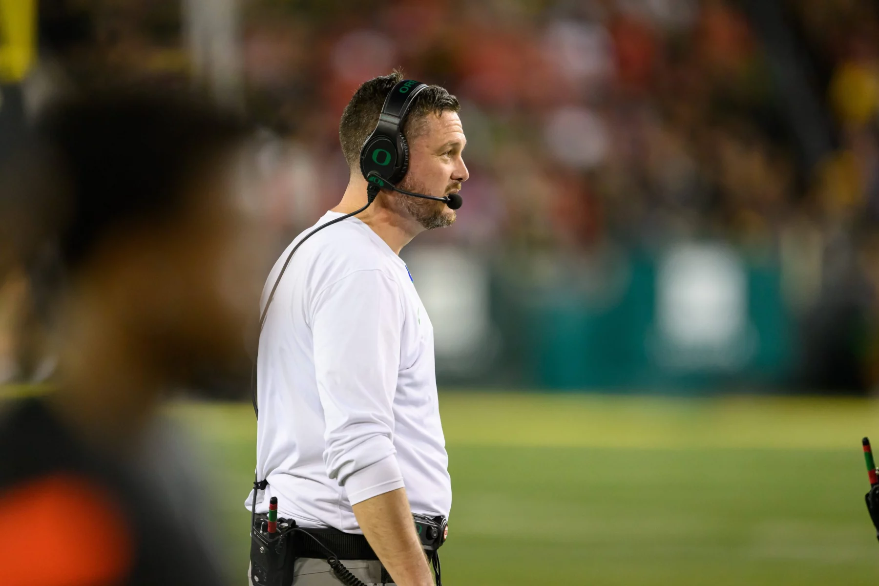 FootballR - NCAA College Football - Ein Trainer, Dan Lanning, in einem weißen Langarmshirt mit Kopfhörern steht am Spielfeldrand eines NCAA-Showdowns. Die Spieler und das Publikum von Ohio State sind im verschwommenen Hintergrund leicht zu erkennen. Die Szene fängt die Intensität dieses nächtlichen Spiels in Oregon ein und hebt das konzentrierte Profil des Trainers hervor. Diese Beschreibung wurde automatisch generiert.