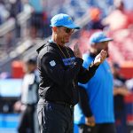 FootballR - NFL - Ein Mann in einem schwarzen Kapuzenpullover und einer blauen Kappe, die an Jim Harbaugh typischen Stil erinnert, trägt eine Sonnenbrille und applaudiert am Spielfeldrand. Eine weitere Person in ähnlicher Kleidung steht in der Nähe. Der Hintergrund ist verschwommen und zeigt Stadionsitze und Zuschauer. Diese Beschreibung wurde automatisch generiert.
