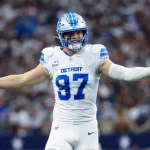 FootballR - NFL - Aidan Hutchinson in der weißen Uniform der Detroit Lions mit der Nummer 97 steht mit ausgestreckten Armen auf dem Feld, als würde er ein automatisch ablaufendes Spiel umarmen. Er trägt einen blauen Helm mit einem Löwenlogo, während die verschwommene Menge im Stadion aufmerksam zusieht. Sein Gesichtsausdruck ist engagiert und fragend zugleich. Er trägt weiße Handschuhe. Diese Beschreibung wurde automatisch generiert.