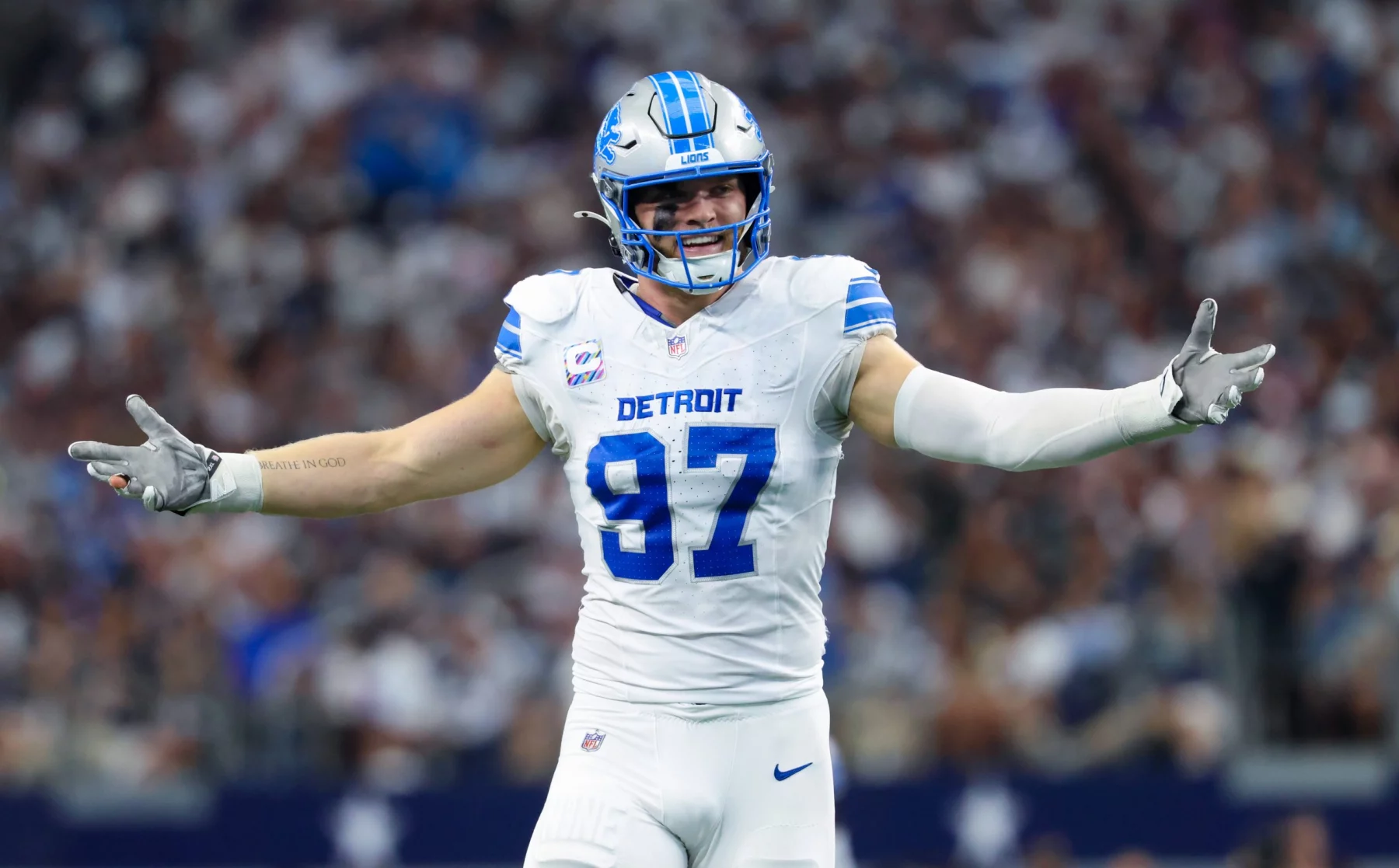 FootballR - NFL - Aidan Hutchinson in der weißen Uniform der Detroit Lions mit der Nummer 97 steht mit ausgestreckten Armen auf dem Feld, als würde er ein automatisch ablaufendes Spiel umarmen. Er trägt einen blauen Helm mit einem Löwenlogo, während die verschwommene Menge im Stadion aufmerksam zusieht. Sein Gesichtsausdruck ist engagiert und fragend zugleich. Er trägt weiße Handschuhe. Diese Beschreibung wurde automatisch generiert.
