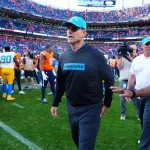 FootballR - NFL - Ein Trainer in einem Chargers-Kapuzenpulli und einer Kappe läuft über ein Footballfeld. Im Hintergrund versammeln sich Spieler, darunter einige in orange-blauen Broncos-Uniformen. Ein Schiedsrichter und ein weiterer Trainer sind in der Nähe, während die Zuschauer die Sitze im Stadion füllen. Jim Harbaugh's Anwesenheit verleiht der Szene Spannung. Diese Beschreibung wurde automatisch generiert.