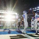 FootballR - NFL Week 6 - Footballspieler in weißen Uniformen feiern in Woche 6 der NFL einen Touchdown und geben sich in der Nähe der Endzone des Stadions High Fives. Sonnenlicht fällt durch große Fenster und sorgt für ein helles Licht. Im Hintergrund sind Spieler der gegnerischen Mannschaft und Schiedsrichter zwischen leuchtend blauen Spielfeldmarkierungen zu sehen. Diese Beschreibung wurde automatisch generiert.