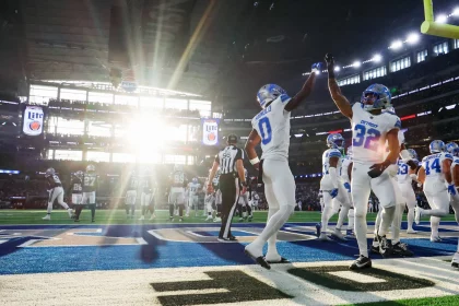 FootballR - NFL Week 6 - Footballspieler in weißen Uniformen feiern in Woche 6 der NFL einen Touchdown und geben sich in der Nähe der Endzone des Stadions High Fives. Sonnenlicht fällt durch große Fenster und sorgt für ein helles Licht. Im Hintergrund sind Spieler der gegnerischen Mannschaft und Schiedsrichter zwischen leuchtend blauen Spielfeldmarkierungen zu sehen. Diese Beschreibung wurde automatisch generiert.