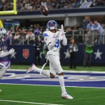 FootballR - NFL - Jameson Williams von den Lions in weiß-blauer Uniform bereitet sich während eines Spiels darauf vor, einen Pass zu fangen. Er steht auf einem grünen Feld mit einem Sternlogo, während ein anderer Spieler in Marineblau hinter ihm herläuft. Im Hintergrund sind eine Menge Zuschauer und ein Logo der Heilsarmee an der Wand zu sehen. Diese Beschreibung wurde automatisch generiert.