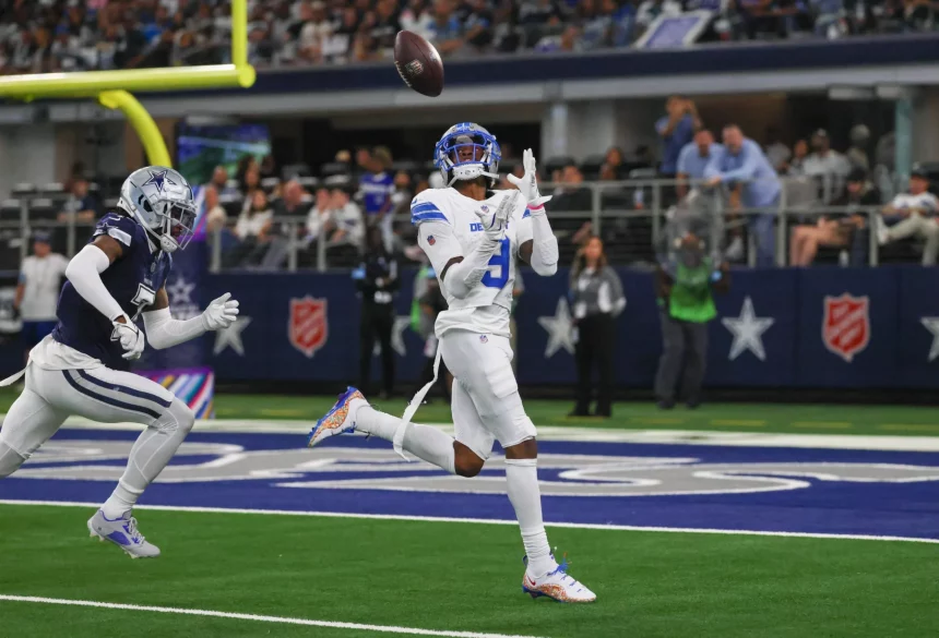 FootballR - NFL - Jameson Williams von den Lions in weiß-blauer Uniform bereitet sich während eines Spiels darauf vor, einen Pass zu fangen. Er steht auf einem grünen Feld mit einem Sternlogo, während ein anderer Spieler in Marineblau hinter ihm herläuft. Im Hintergrund sind eine Menge Zuschauer und ein Logo der Heilsarmee an der Wand zu sehen. Diese Beschreibung wurde automatisch generiert.