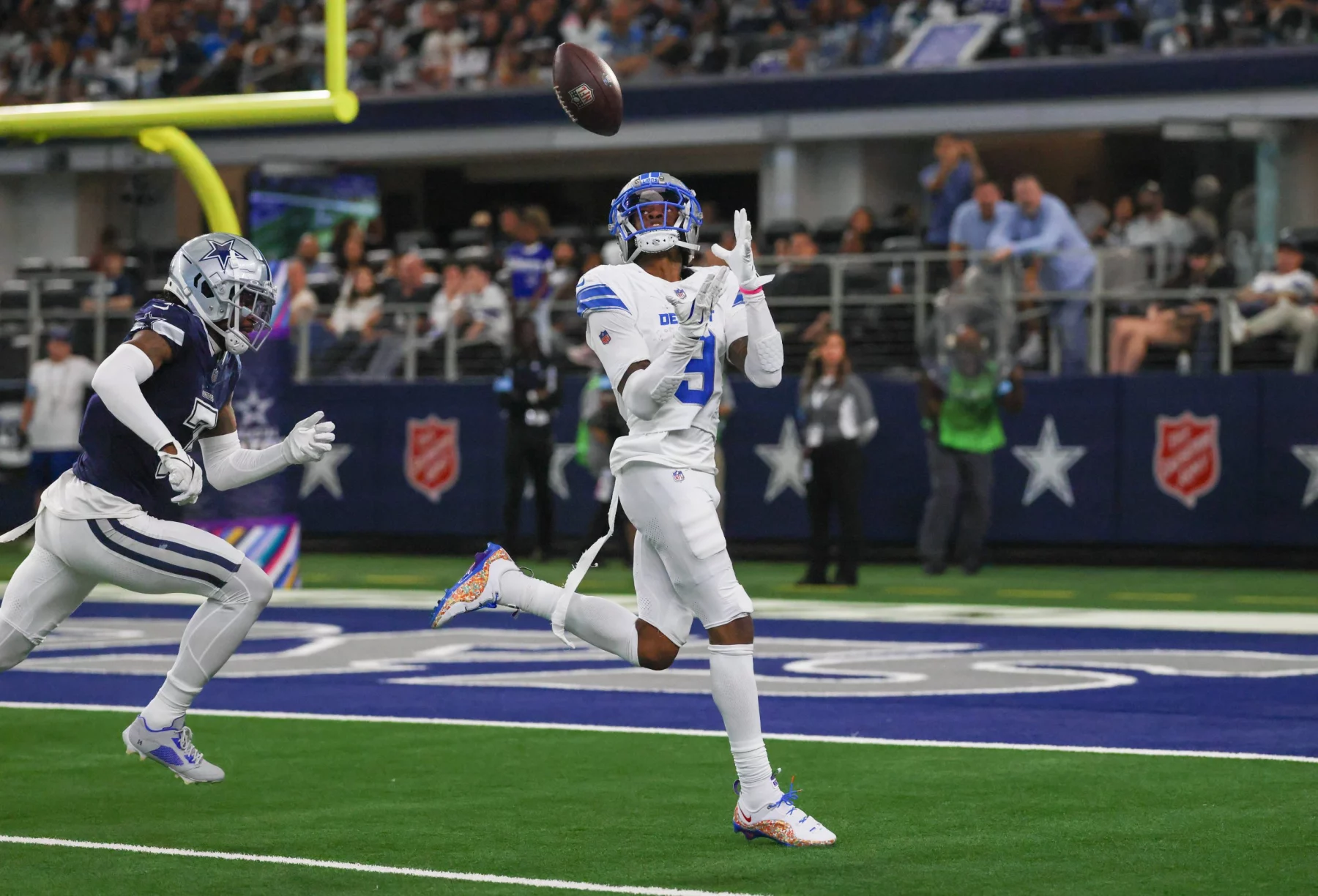 FootballR - NFL - Jameson Williams von den Lions in weiß-blauer Uniform bereitet sich während eines Spiels darauf vor, einen Pass zu fangen. Er steht auf einem grünen Feld mit einem Sternlogo, während ein anderer Spieler in Marineblau hinter ihm herläuft. Im Hintergrund sind eine Menge Zuschauer und ein Logo der Heilsarmee an der Wand zu sehen. Diese Beschreibung wurde automatisch generiert.